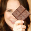 A close-up of a woman holding a square dark chocolate bar with four embossed unicorn motifs, with her face softly blurred in the background.