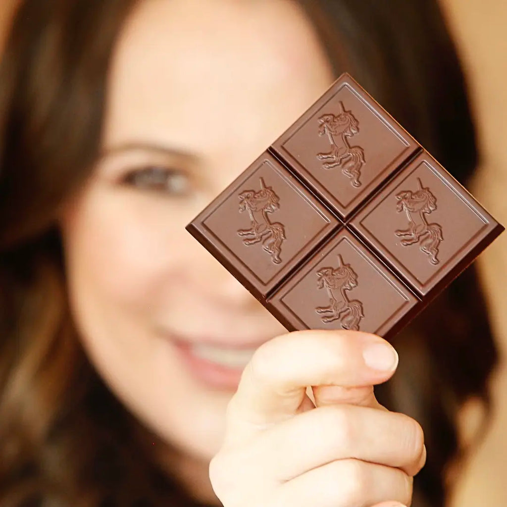 A close-up of a woman holding a square dark chocolate bar with four embossed unicorn motifs, with her face softly blurred in the background.