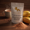 A stand-up pouch of Addictive Wellness Wild Cacao Powder stands on a wooden surface with cocoa powder spilled in front, a mug of hot cocoa with steam in the background, and cacao beans nearby. The pouch is white with a gold unicorn logo and gold text.