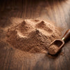 A mound of fine brown powder, likely cocoa powder or coffee grounds, scattered on a dark wooden surface next to a small wooden scoop with a brass-colored accent.