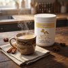 A white plastic tub of Addictive Wellness Elixir Blends Focused Energy Chai sits on a rustic wooden table next to a ceramic mug of steaming chai, with cinnamon sticks and star anise scattered around.