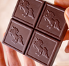 A close-up of a dark chocolate bar held in hands, showing four square sections, each embossed with a unicorn design.