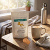 A stand-up pouch of Cordyceps Premix Power supplement by Addictive Wellness sits on a rustic wooden table beside a cup of coffee, a spoon, and a small pile of the tan powder. A silver water bottle stands near a window in the softly lit background.
