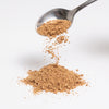 A silver spoon holding a small amount of fine brown powder above a pile of the same powder on a clean white background.