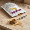 A white stand-up pouch labeled 'Doctor Wellness Mangosteen' with a gold horse logo and purple band, shown on a beige stone surface with a metal spoon holding loose brown powder spilled in front.