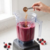 A clear blender pitcher filled with a deep berry smoothie sits on a blender base in a bright kitchen, while a hand adds a brown powder from a metal measuring spoon.