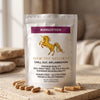 A stand-up pouch of Addictive Wellness Mangosteen capsules sits on a stone surface with several capsules scattered in front. The pouch is white with a purple label band, a gold unicorn graphic, and black text.