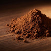 A small mound of fine brown powder, likely cocoa powder or ground coffee, scattered on a dark wooden surface with a warm, moody background.