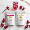 Two stand-up pouches of Addictive Wellness herbal supplements on a white wooden surface, surrounded by red dragon fruit and a glass of red drink with ice. The left pouch is labeled Robelle Hibiscus with a pink label, and the right pouch is labeled Astragalus with a yellow label.