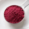 A metal measuring scoop filled with a vivid deep pinkish-red powder, photographed from above on a clean white background.