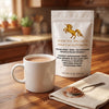 A stand-up pouch of Addictive Wellness Wild Cacao Powder stands on a wooden table beside a white mug of hot cocoa, with a spoon of powder and a folded cloth in a warm kitchen setting.