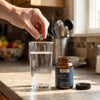 Hand about to drop a small gray H2 Elite tablet into a glass of water beside the jar