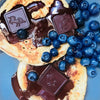 Two golden pancakes topped with glossy chocolate syrup, dark chocolate squares embossed with a unicorn, and fresh blueberries on a blue plate.