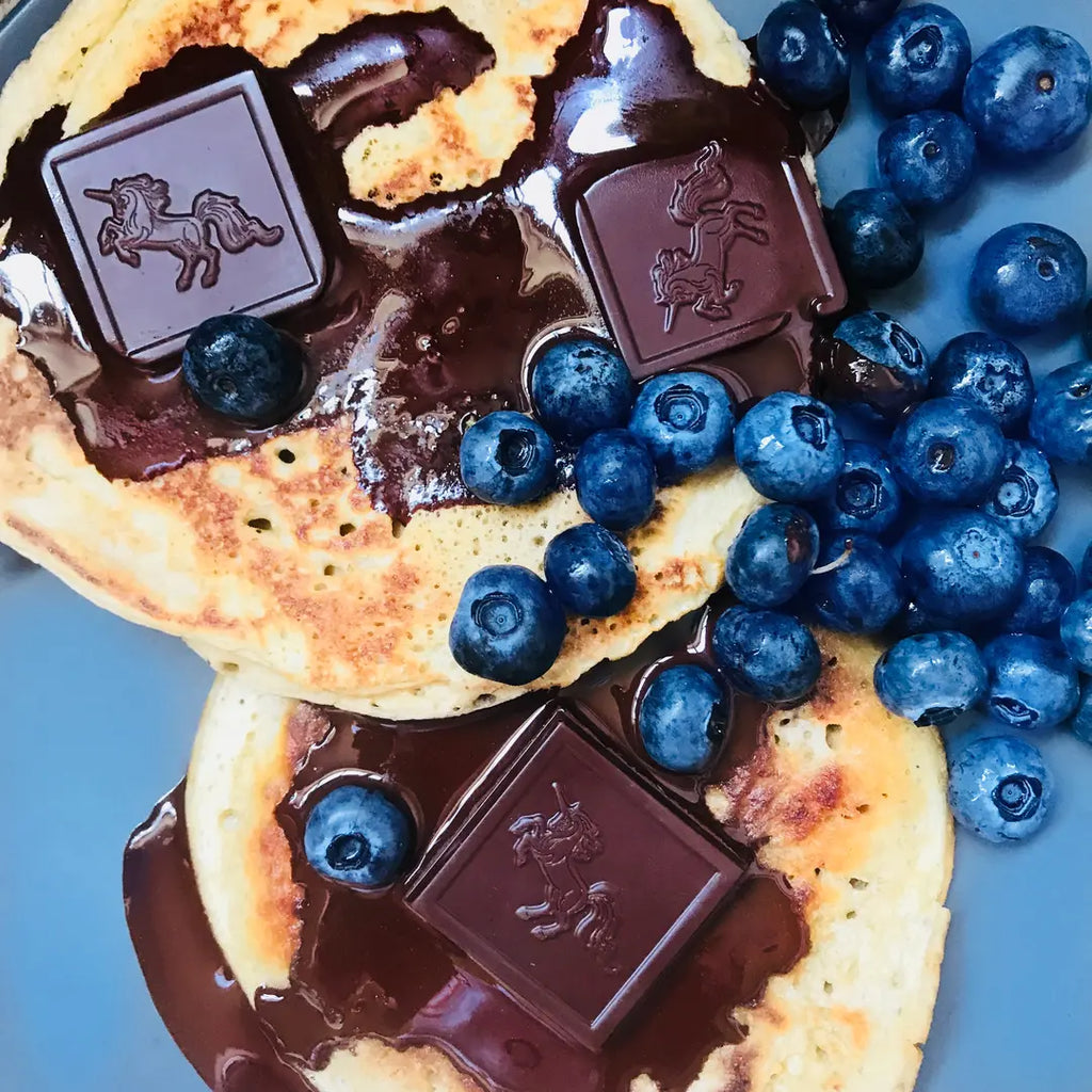 Two golden pancakes topped with glossy chocolate syrup, dark chocolate squares embossed with a unicorn, and fresh blueberries on a blue plate.