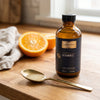 One teaspoon of Liposomal Vitamin C in a brass spoon with the bottle and a halved orange on a kitchen counter